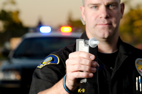 A police officer holds a breath test machine in his hand ready at a traffic stop with his patrol car in the background.